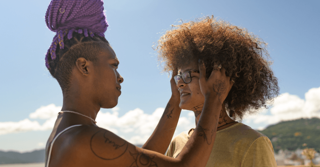 Existir fora do binário: Duas pessoas negras e não binárias interagindo, onde uma arruma o óculos da outra, se olhando com semblante feliz e seguro.