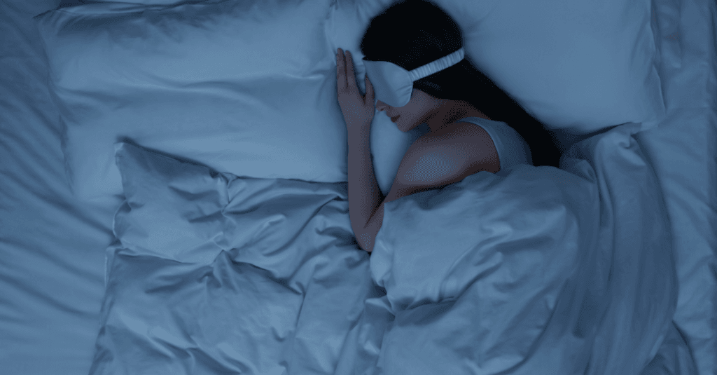 Uma pessoa jovem adulta aconchegada em uma cama usando um cobertor pesado (weighted blanket) e uma máscara de dormir, com o quarto em meia-luz suave.