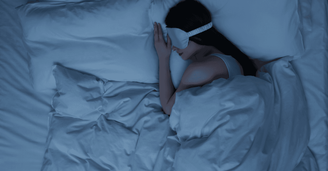 Uma pessoa jovem adulta aconchegada em uma cama usando um cobertor pesado (weighted blanket) e uma máscara de dormir, com o quarto em meia-luz suave.